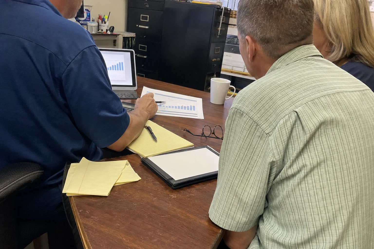 Agent in a navy polo points a pen at a printed bar chart on a wooden desk as a man and woman look on, in a cluttered back office with black filing cabinets