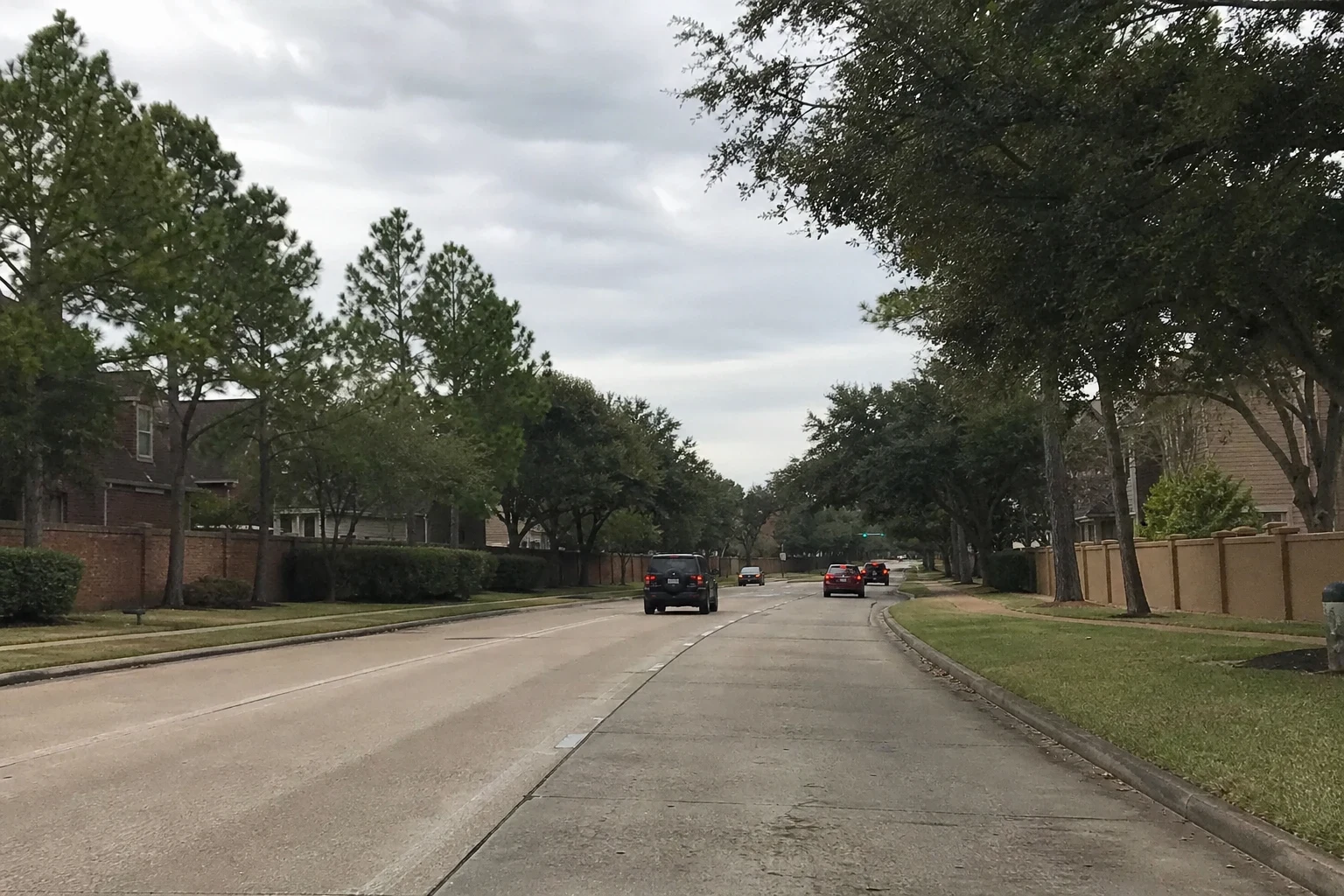 Tree-lined suburban concrete road under an overcast sky, with brick and stucco perimeter walls, grassy medians and a few cars driving ahead
